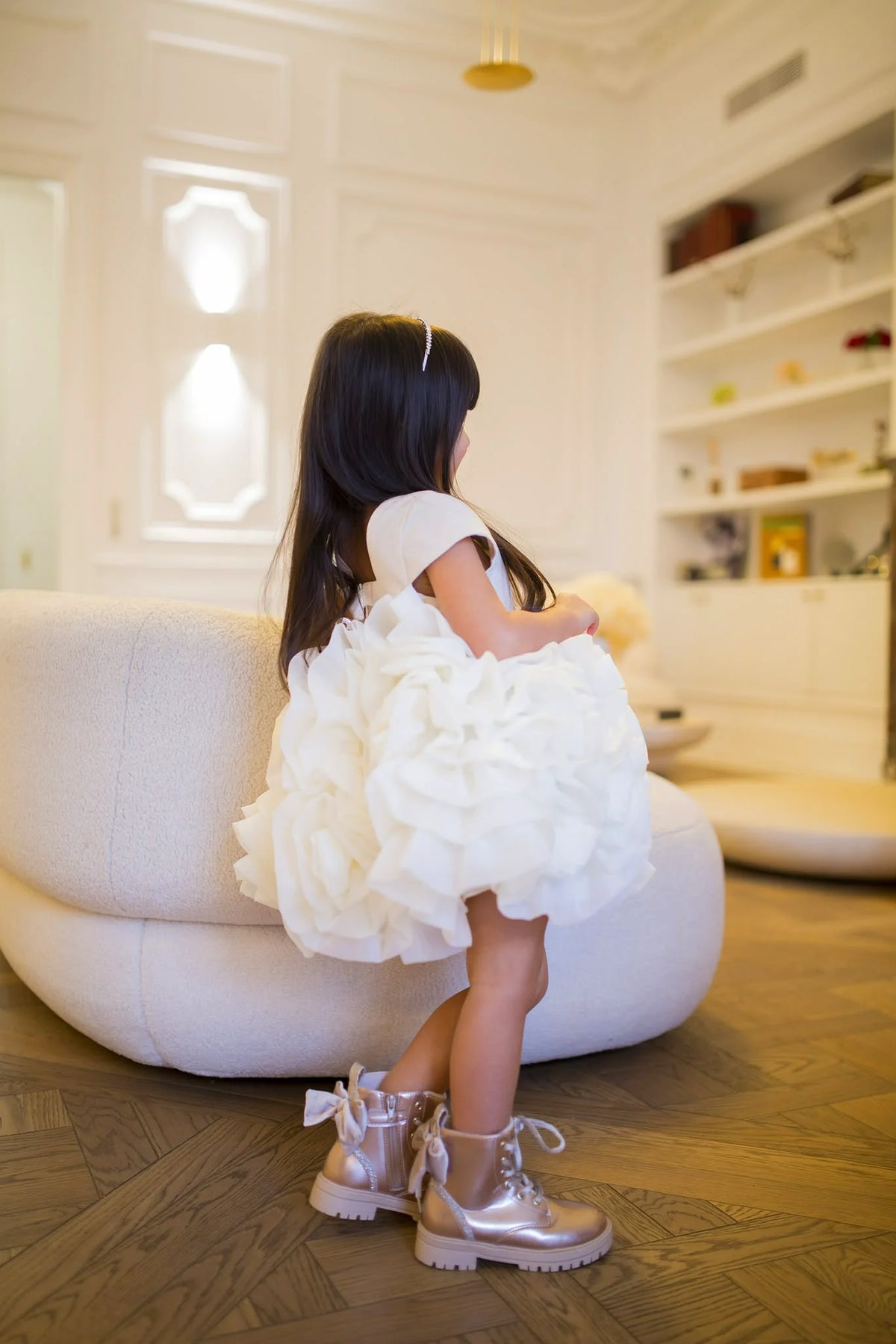 Enfant assis sur un fauteuil blanc, portant une robe blanche texturée et des bottines métalliques dans un intérieur lumineux. Image