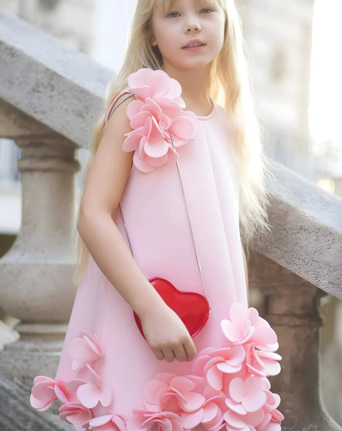 Jeune enfant portant une robe rose pâle ornée d’applications florales sur un escalier en pierre.